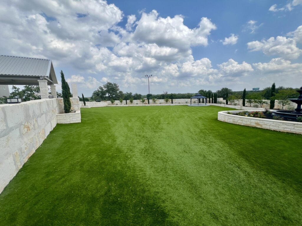 Open grassy courtyard with stone walls, a covered pavilion, and a fountain surrounded by landscaping under a partly cloudy sky