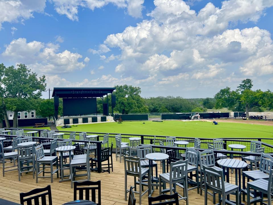 Round Mountain Cider Mill outdoor concert stage and seating area in Texas, featuring a large lawn and elevated patio tables under a blue sky