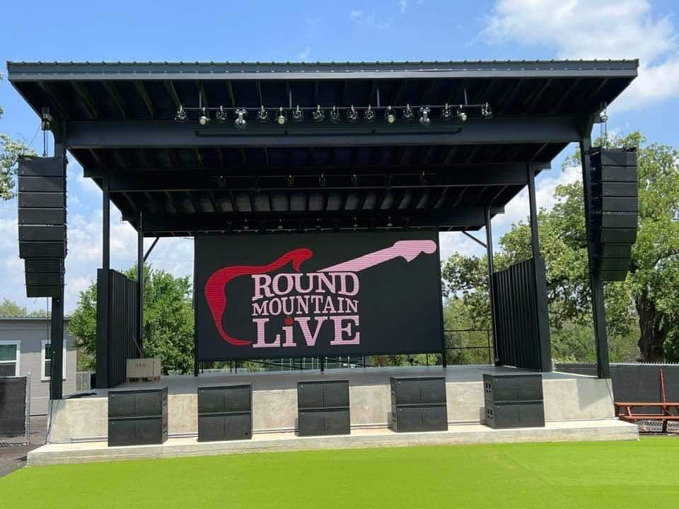 Outdoor concert stage at Round Mountain Live in Texas featuring large speakers and LED screen with guitar logo on artificial turf
