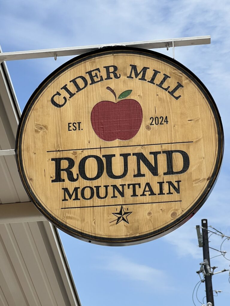 Round wooden sign for Round Mountain Cider Mill in Texas, featuring a red apple graphic and "Est. 2024" under a blue sky