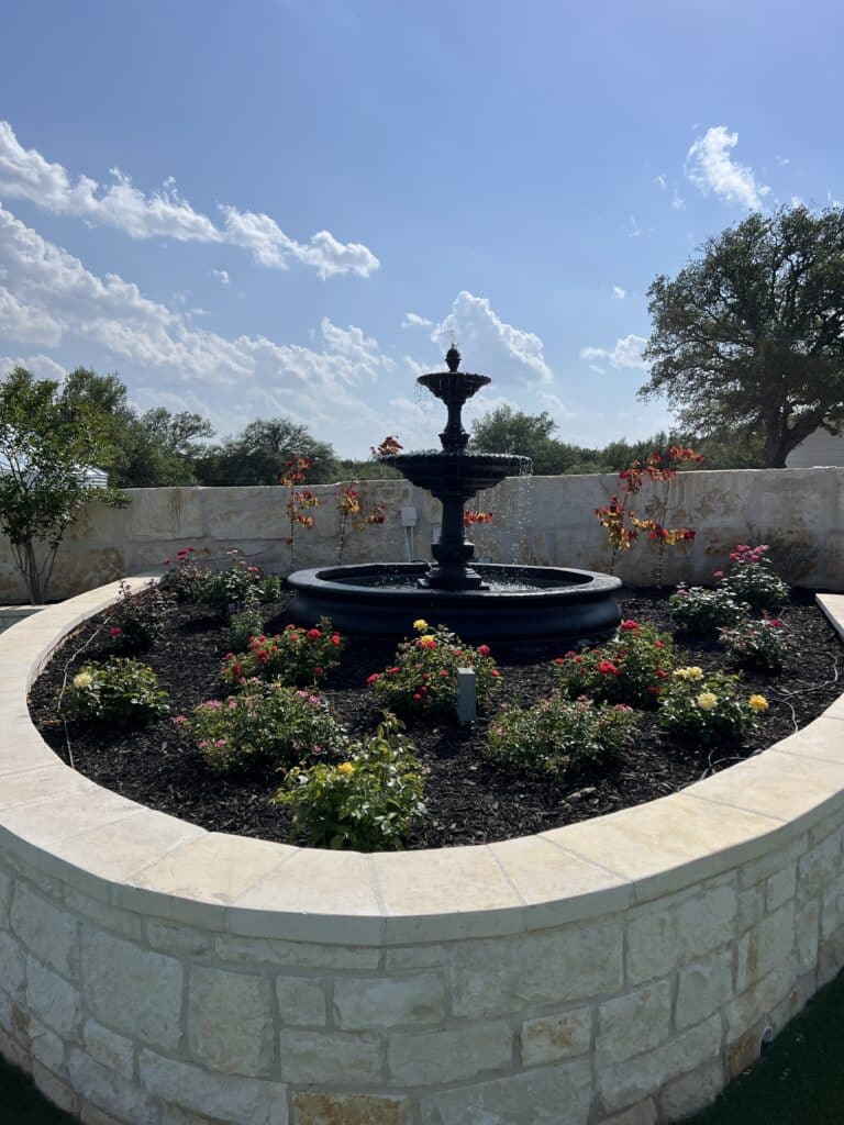 Tiered black fountain surrounded by blooming rose bushes and stone wall garden at Round Mountain Cider Mill in Texas