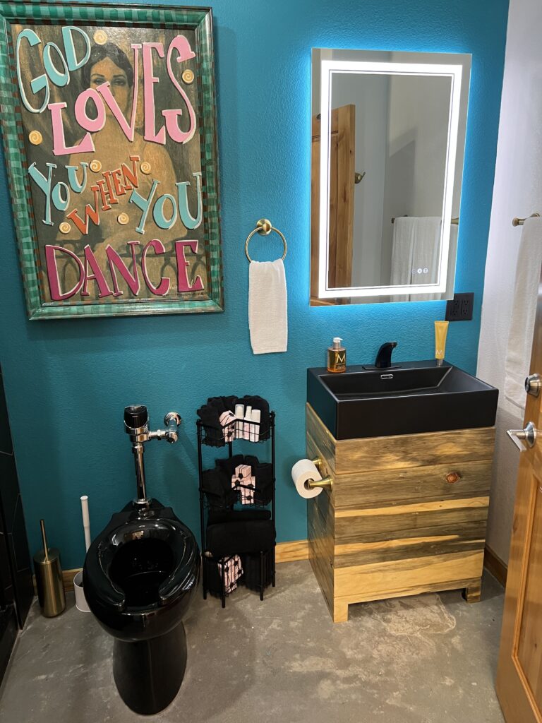 Colorful bathroom at Round Mountain Cider Mill in Texas featuring a black toilet, rustic wood vanity with black sink, and framed wall art reading “God loves you when you dance”