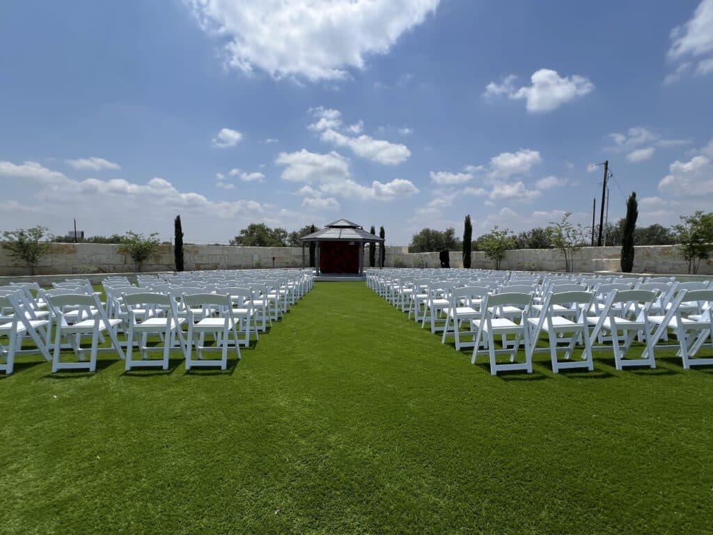 Outdoor wedding ceremony setup at Round Mountain Cider Mill in Texas with white folding chairs facing a gazebo and rose backdrop on green turf