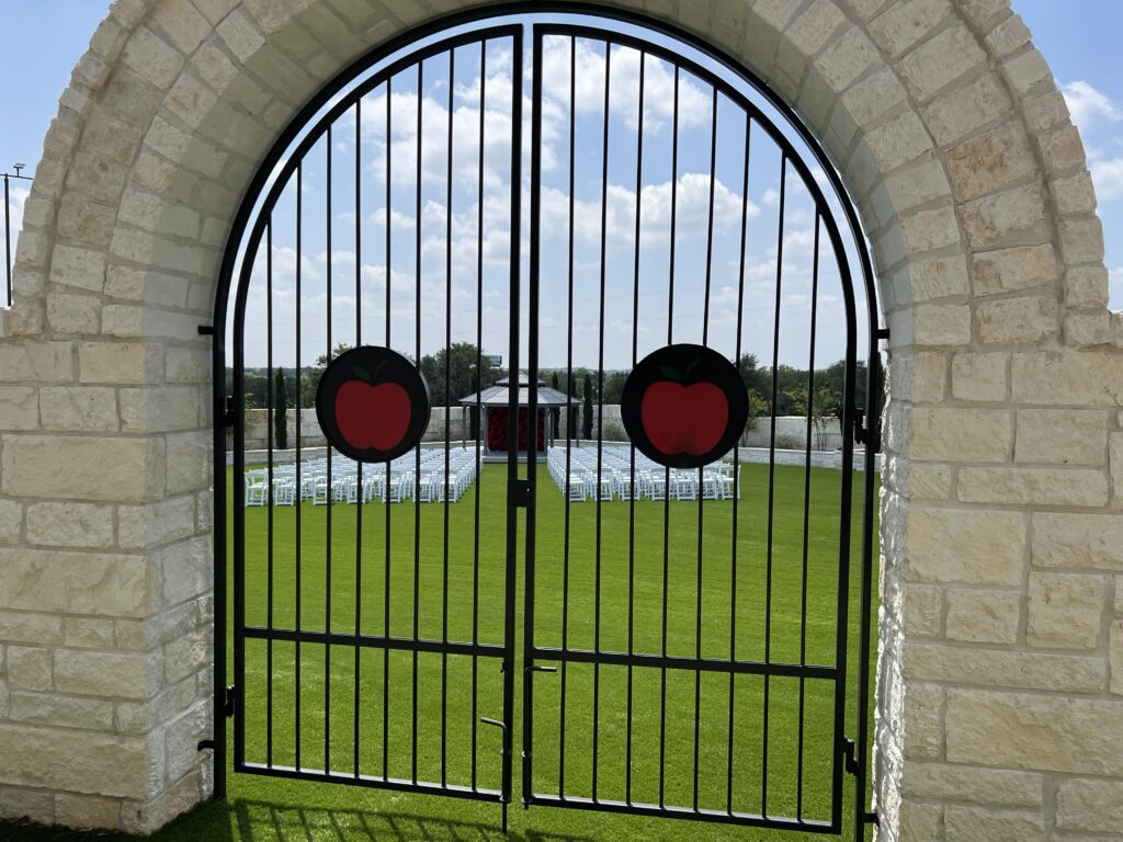 Wrought iron gate with red apple emblems framing the outdoor wedding lawn at Round Mountain Cider Mill in Texas, with rows of white chairs and gazebo visible in background
