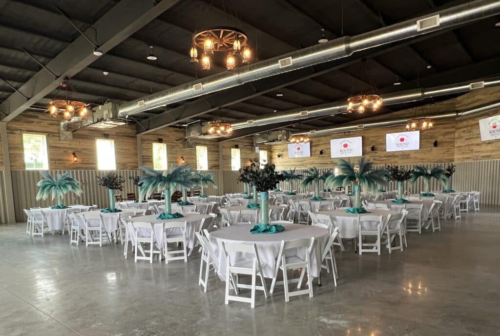 Indoor reception hall at Round Mountain Cider Mill in Texas with round tables, white chairs, teal centerpieces, and industrial rustic décor