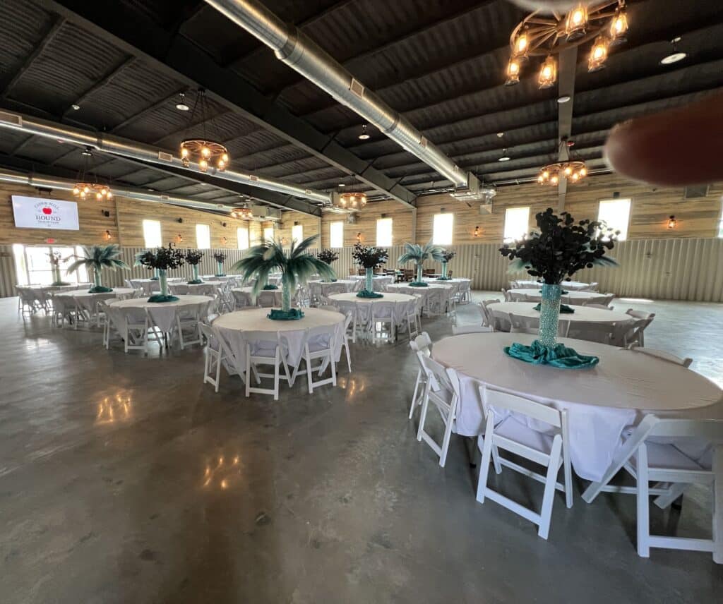 Wide-angle view of decorated indoor event space at Round Mountain Cider Mill in Texas with white round tables, teal floral centerpieces, and rustic-industrial lighting
