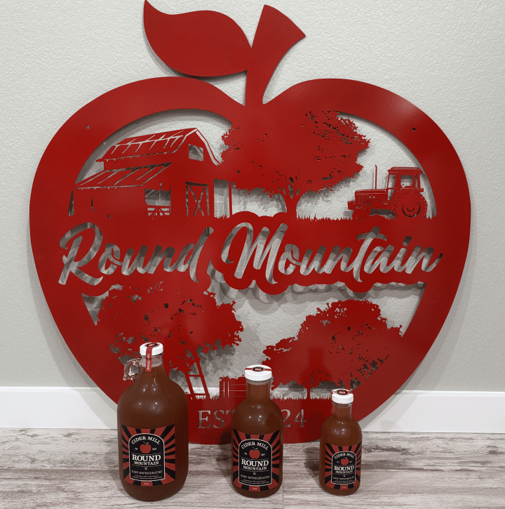 Three bottles of Round Mountain Cider in front of a large red apple-shaped wall sign featuring farm imagery and Round Mountain branding in Texas
