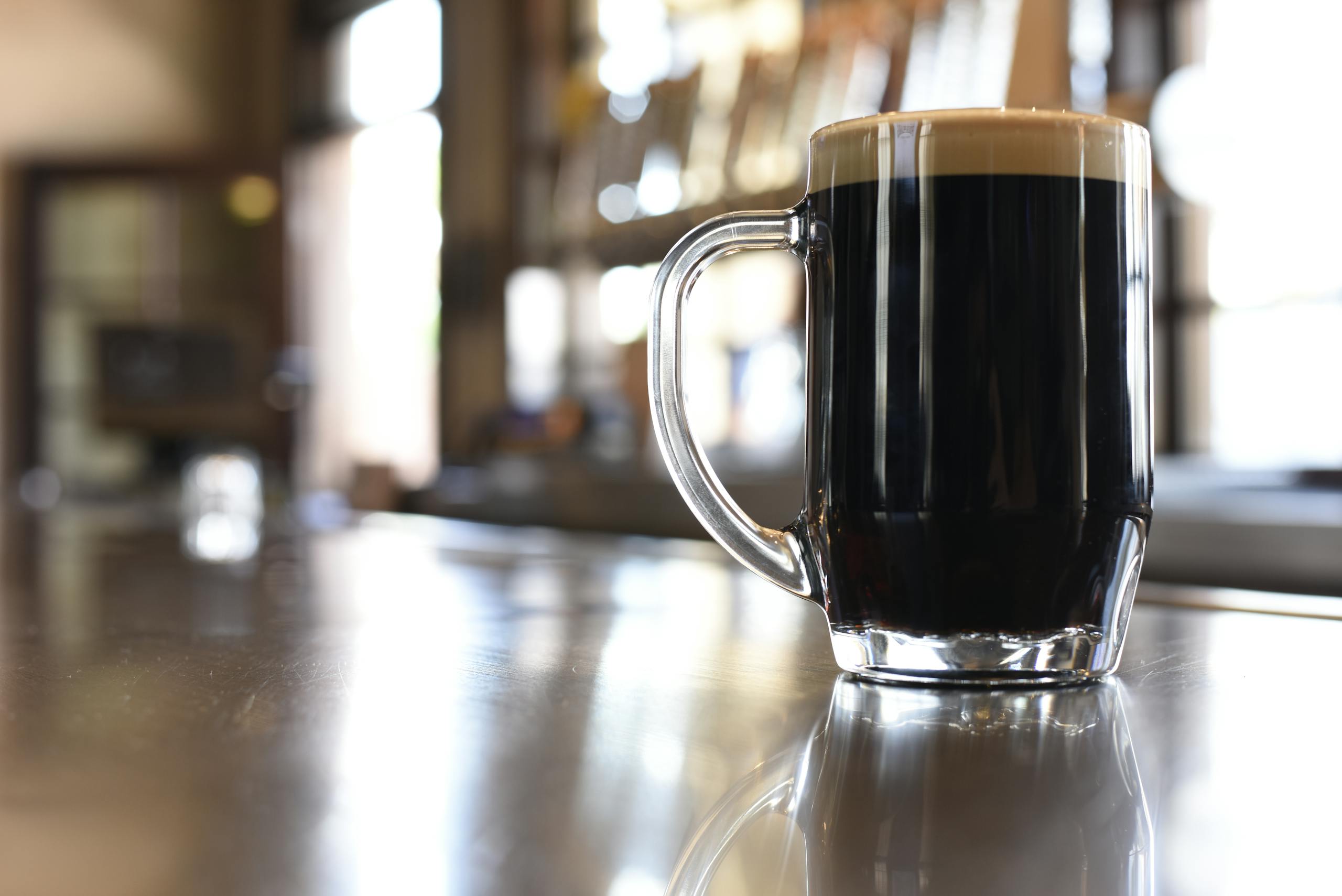 A glass mug of dark beer with foam, set on a bar counter with bokeh background.