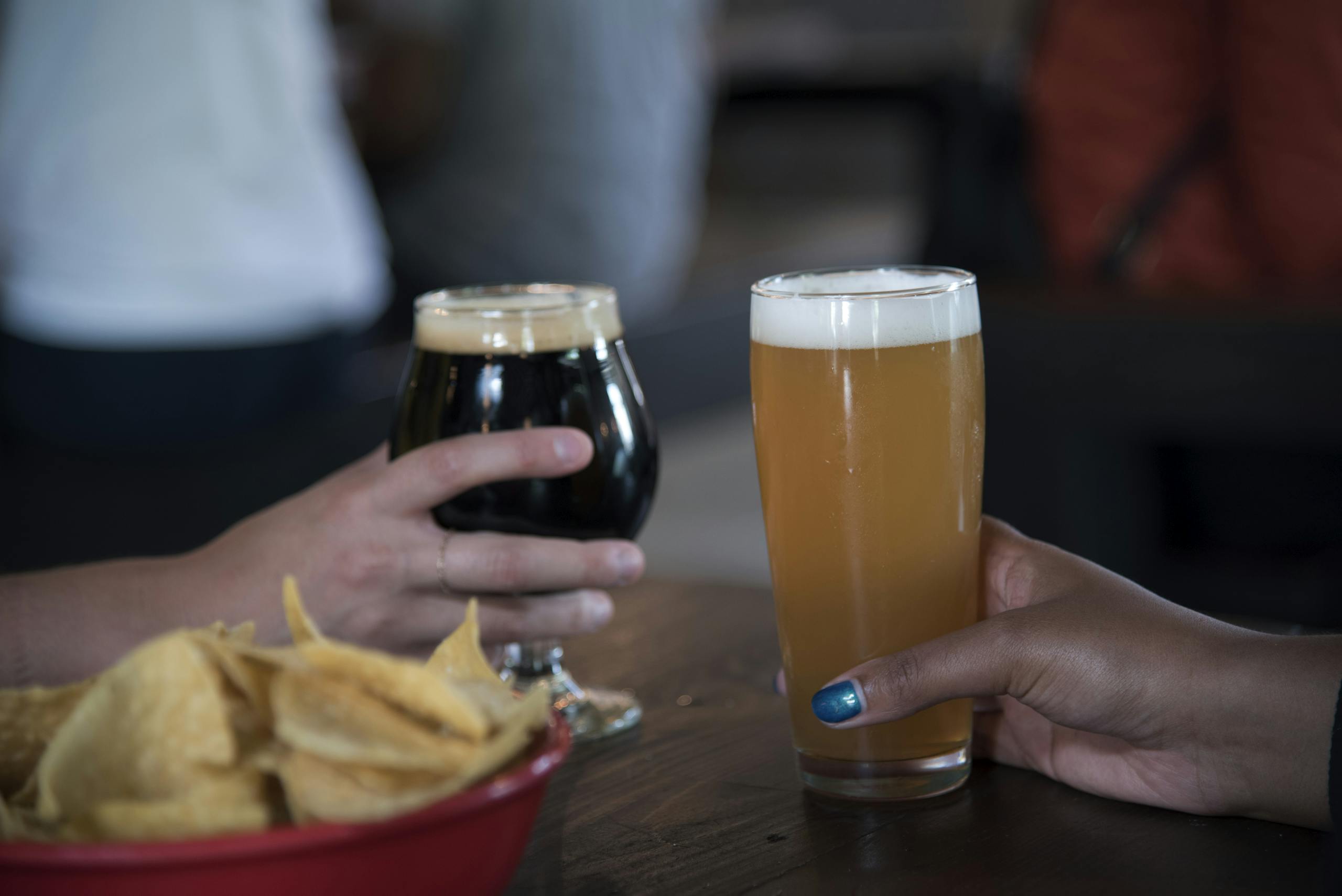 Close-up view of two hands holding beer glasses and a bowl of chips.