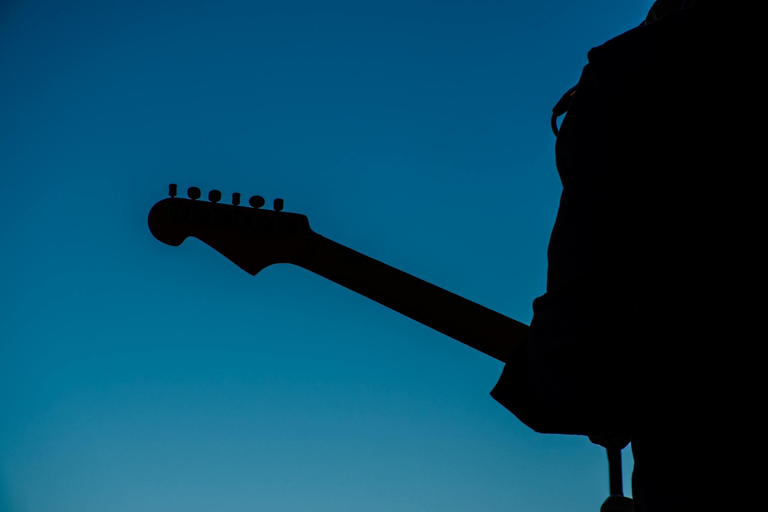 Silhouette of an adult playing an electric guitar against a clear blue sky, creating a dramatic and artistic scene.